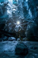 A rugged male explorer looks up at an amazing blue ice cave underneath a glacier.